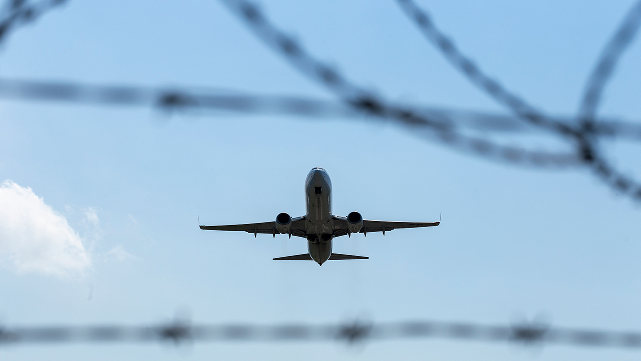 A plane flying in the sky. Barbed wire barrier at the foreground of picrure.